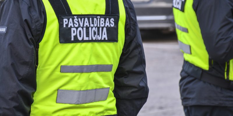 Divi pašvaldības policisti redzami no muguras, tērpušies melnās jakās un spilgti dzeltenās atstarojošās vestēs. Uz kreisā policista vestes ir uzraksts "PAŠVALDĪBAS POLICIJA".