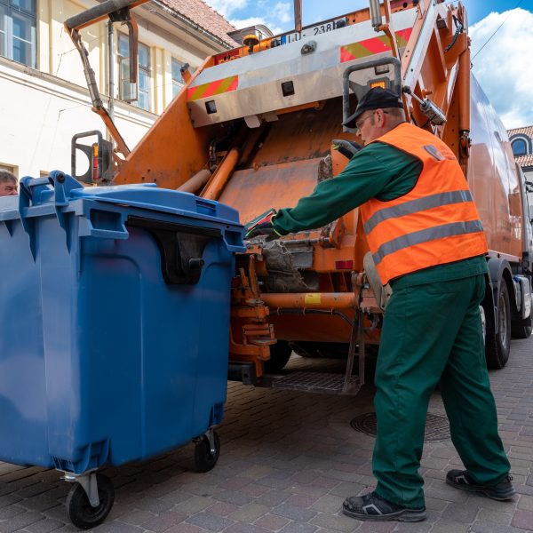 Oranžā atstarojošā vestē tērpies SIA Kuldīgas komunālie sētnieks stumj lielu zilu atkritumu konteineru pie oranžas atkritumu savākšanas mašīnas. Fonā redzamas vecpilsētas ēkas.