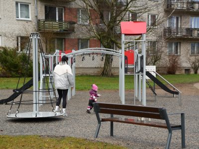 Sieviete sudraba virsjakā un mazs bērns rozā cepurē un violetā kombinezonā atrodas modernā rotaļu laukumā. Sieviete stāv pie rotējoša karuseļa, kamēr bērns iet viņas virzienā. Fonā redzama daudzdzīvokļu māja.