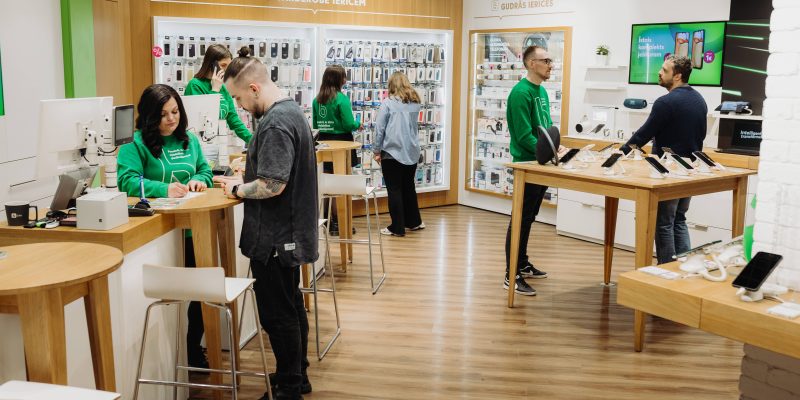 Moderns elektronikas veikals, kurā darbinieki zaļos džemperos apkalpo klientus.