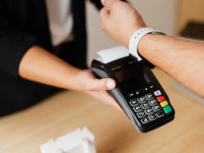 Tuvplānā redzams, kā cilvēks ar baltu viedpulksteni uz rokas veic bezkontakta maksājumu, pieliekot to pie melna POS termināļa, ko tur cits cilvēks.