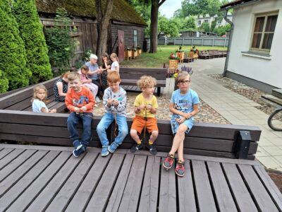 Āra nodarbībā bērnu un pieaugušo grupa veido rokdarbus. Priekšplānā četri zēni sēž uz koka terases, divi no viņiem aktīvi pin krāsainas auklas. Zēnam labajā pusē ir gaiši zils T-krekls ar uzrakstu "SALTY WILD". Fonā redzamas citas personas, vecas koka ēkas un koki.