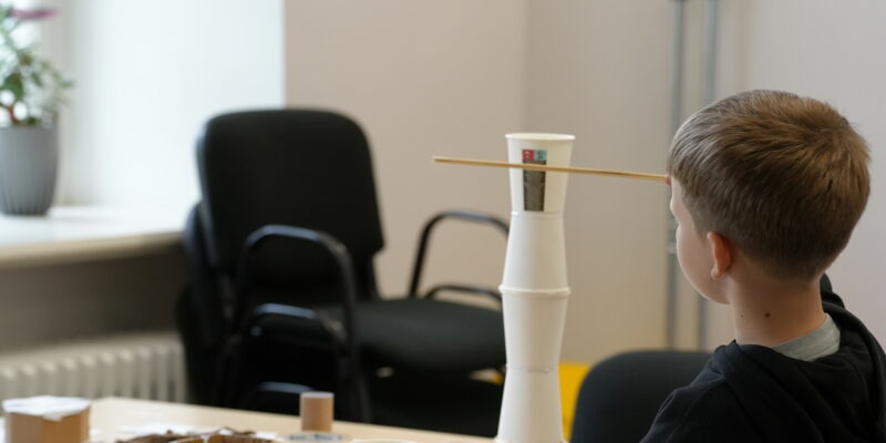 Zēns no muguras stāv pie galda un skatās uz no baltām papīra krūzītēm un koka nūjiņām izveidotu torni. Uz galda redzami citi rokdarbu materiāli.